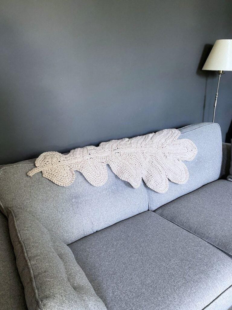 A small grey crochet blanket in the shape of an oak leaf is laying across the back of a grey settee