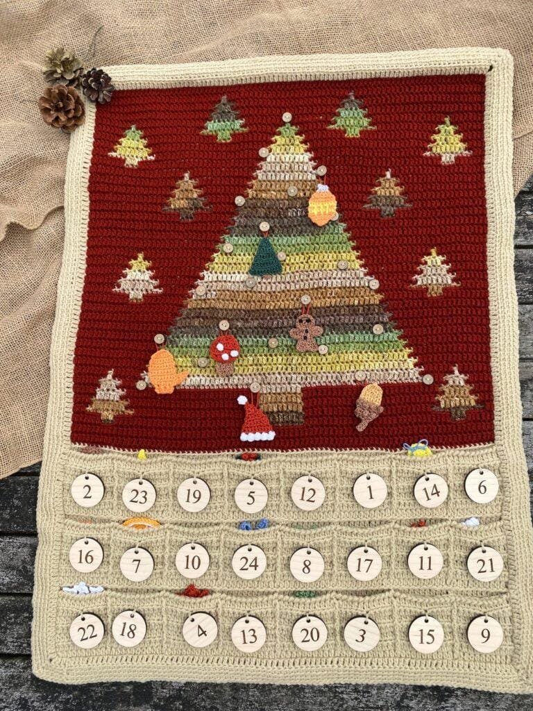 A crochet advent calendar wall hanging shows a green and brown tree on a red background. Pockets underneath with numbers on hold the hanging decorations