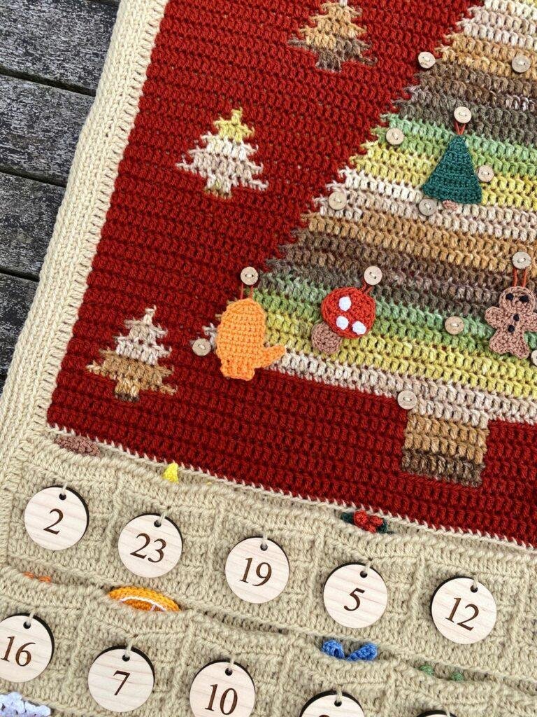 a close up of a crochet advent calendar showing some of the pockets with hanging decorations in and a mitten and toadstool hung on a green and brown tree