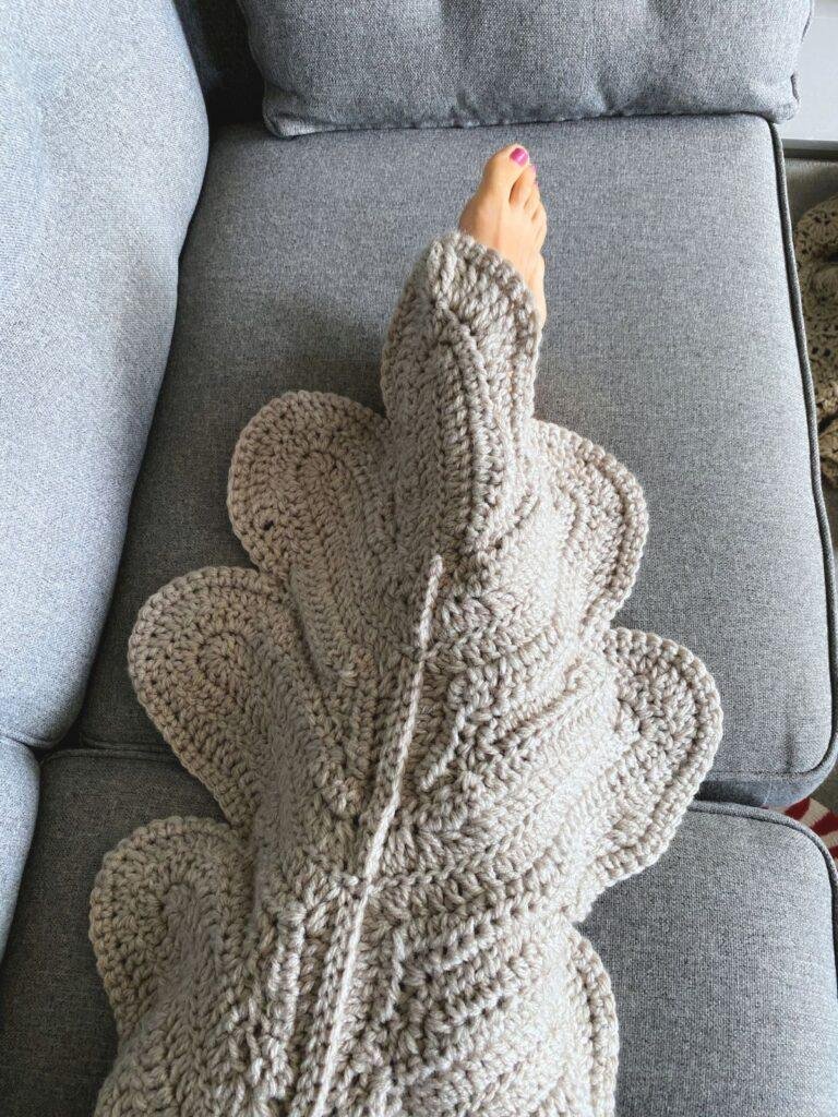 A leaf shaped crochet blanket is laying over someones lap with the toes poking out on a grey sofa