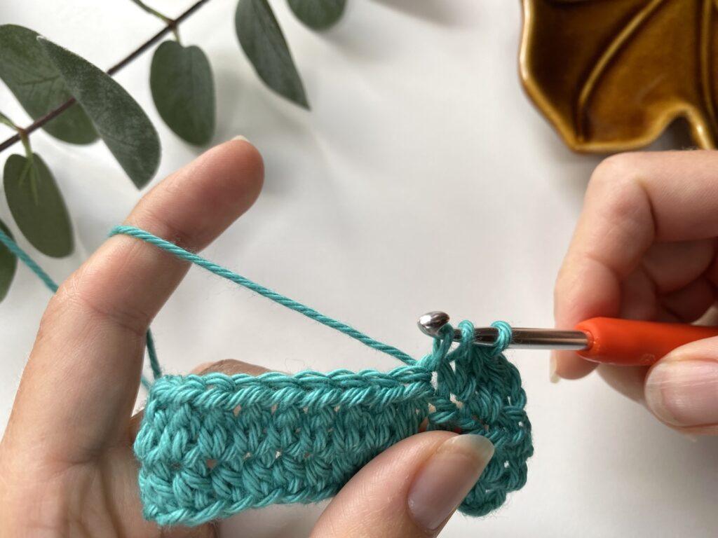 2 hands with crochet hook and teal yarn are making a crochet stitch