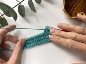 A sample of crochet fabric in teal with 2 hands holding it and some greenery framing the shot