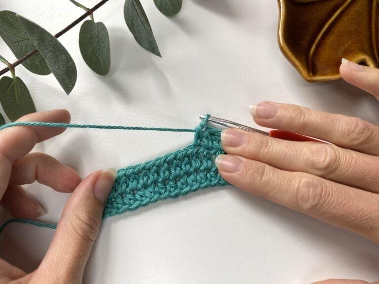 A sample of crochet fabric in teal with 2 hands holding it and some greenery framing the shot
