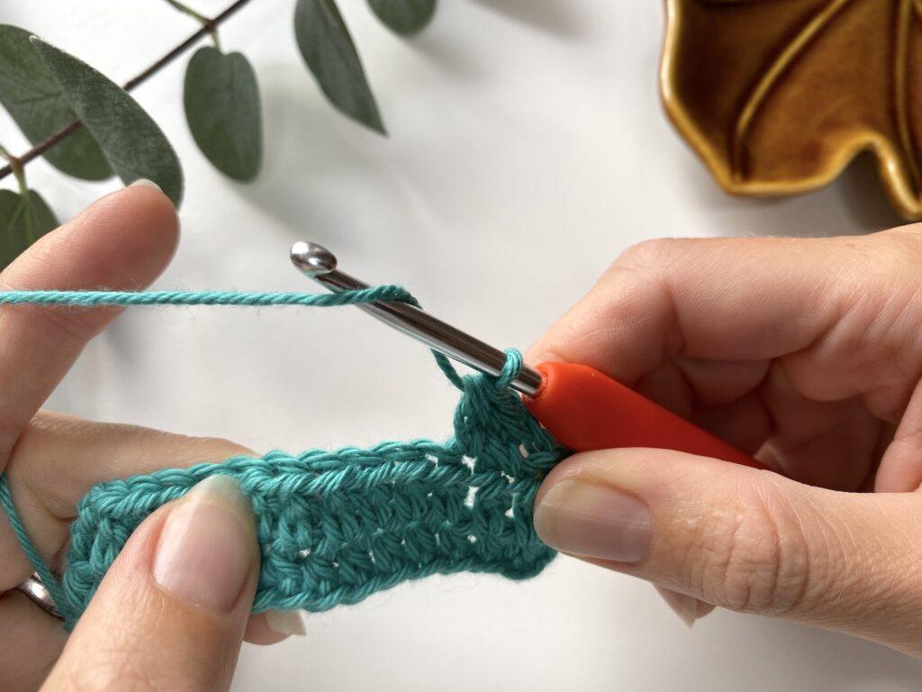 A person holding crochet hook and yarn making a crochet stitch with teal yarn