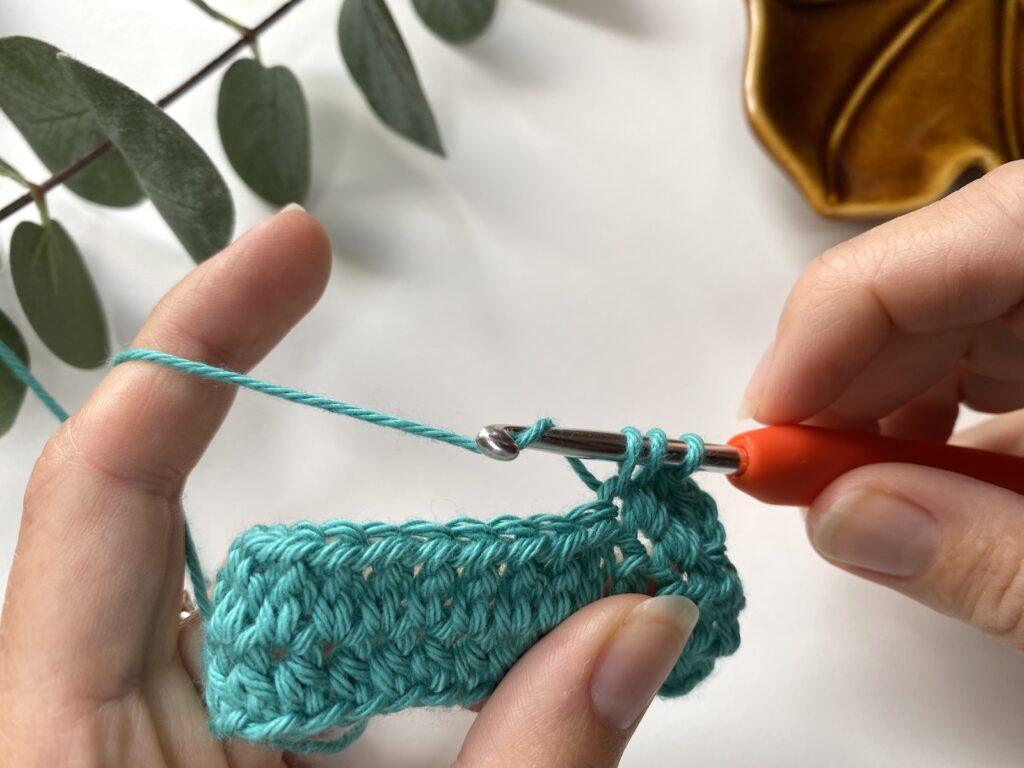 A person partway through making an extended half double crochet stitch with teal yarn
