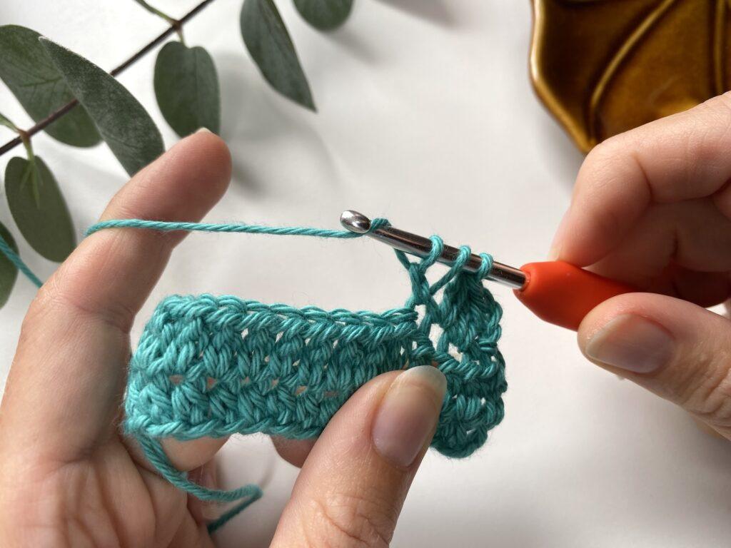 2 hands are holding a crochet fabric and making a stitch with a crochet hook