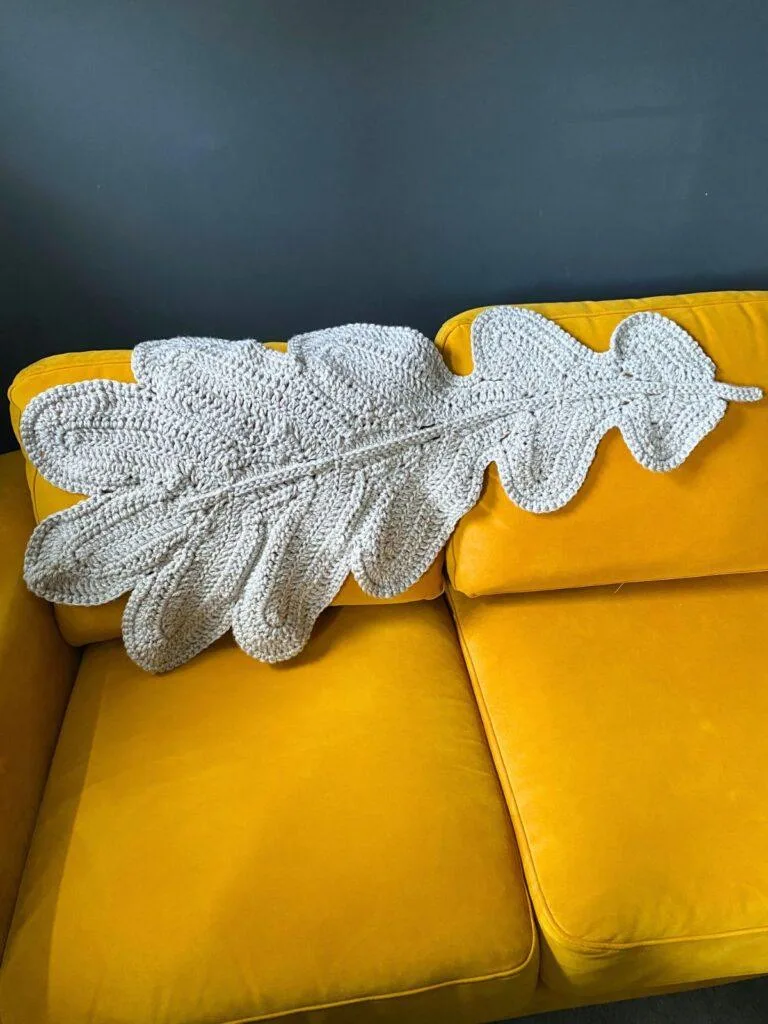 A giant crochet oak leaf in grey is draped over the back of a yellow sofa