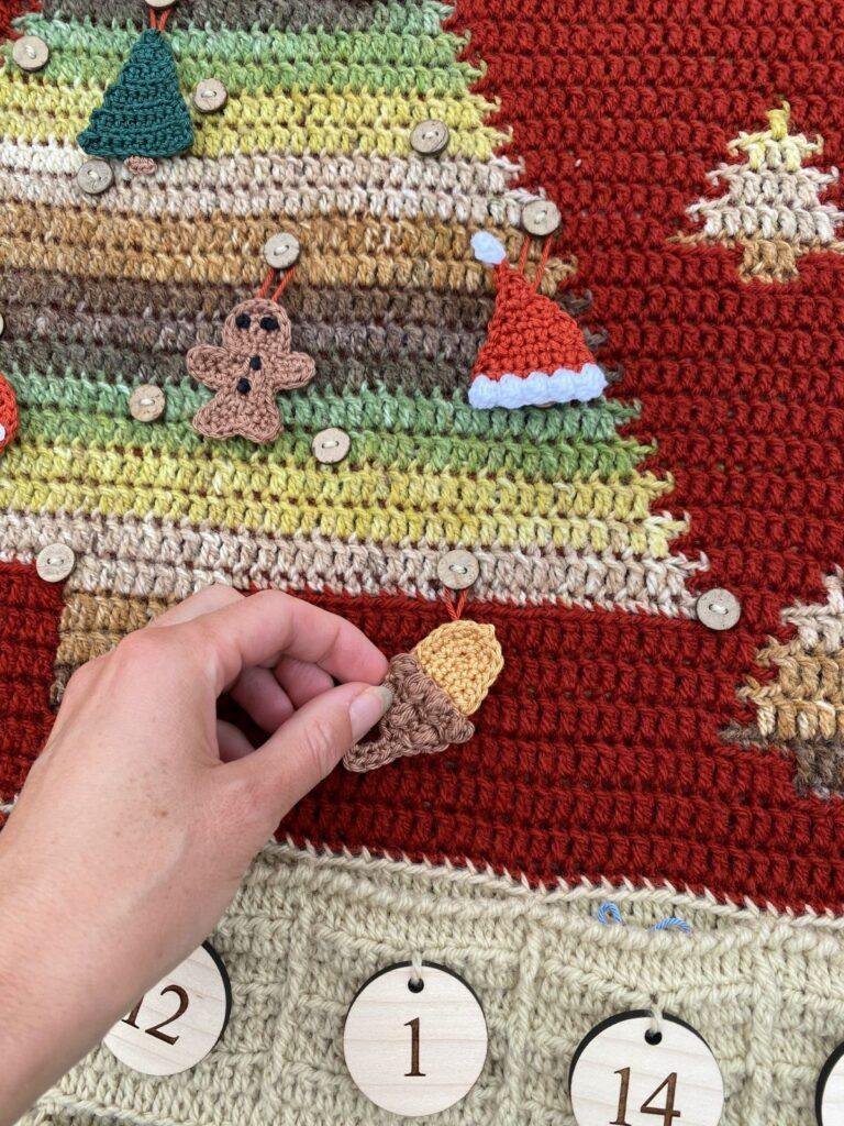 A hand a placing a small crochet acorn decoration onto the crochet advent calendar wall hanging Christmas tree