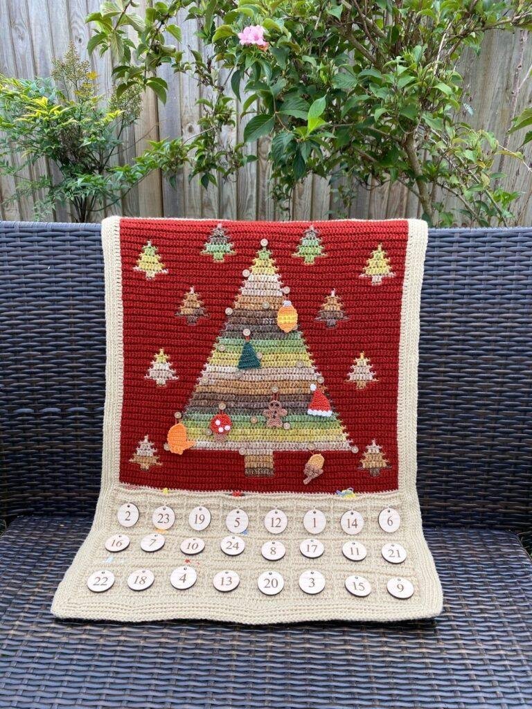 A crochet Christmas tree wall hanging advent calendar lays over a wicker bench in a garden