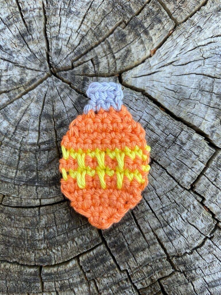 An orange crochet bauble that is flat with yellow zigzags lays on a wooden slice