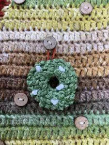 A mini crochet wreath with white berries on is hung on a green and brown crochet background