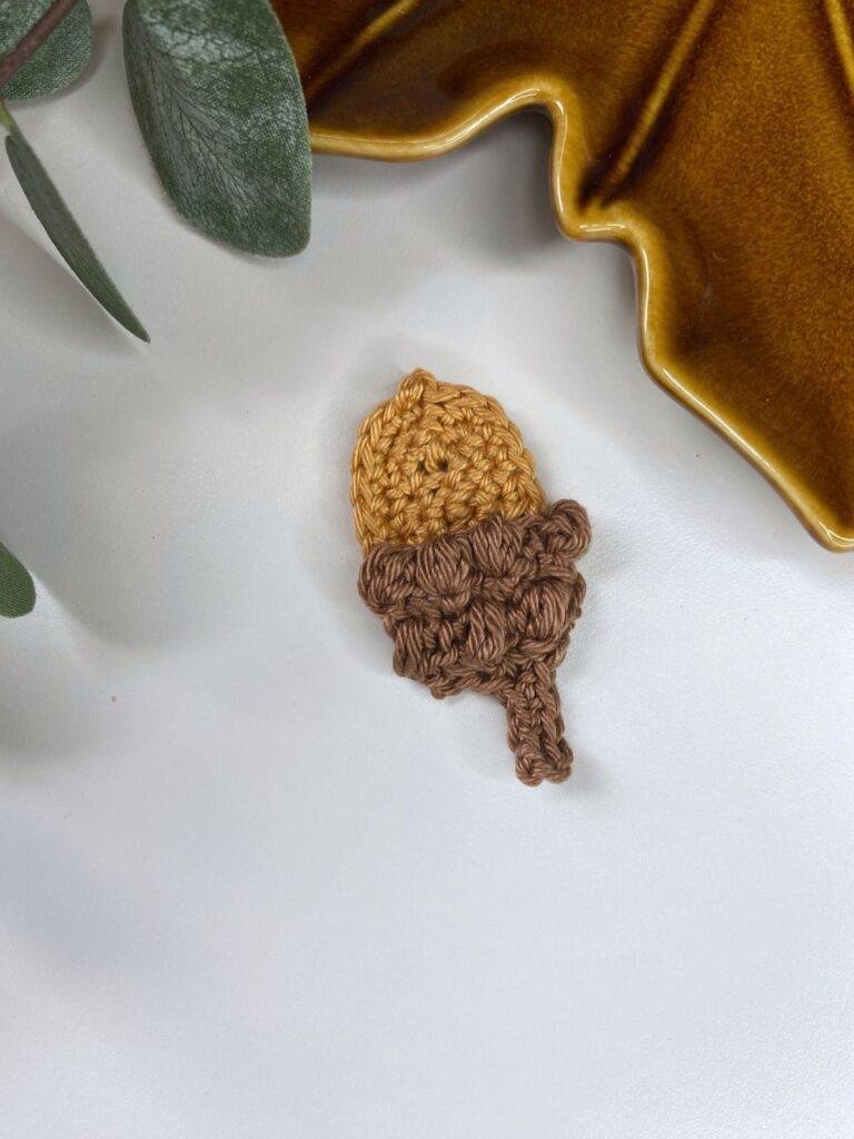 A crochet acorn lays on a white background with a brown leaf shaped dish and a green leaf at the edge