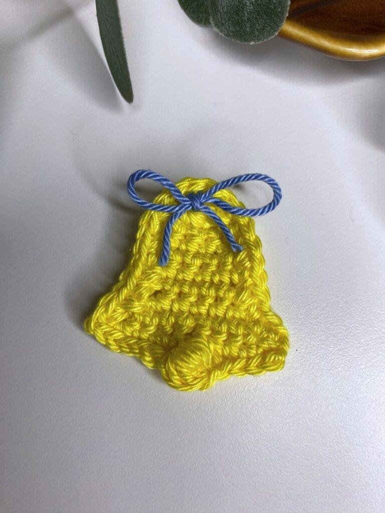 A yellow crochet flat bell on a white background with a brown dish and greenery around