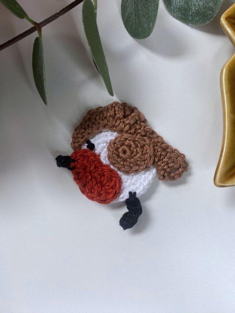 A small flat crochet robin lays on a white background with greenery and the edge of a brown dish showing