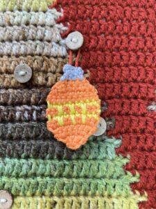 An orange crochet bauble with yellow zigzags hangs on a button on a crochet wall hanging