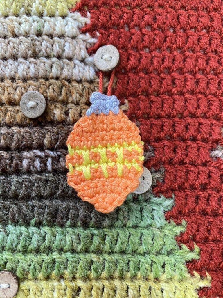 An orange crochet bauble with yellow zigzags hangs on a button on a crochet wall hanging