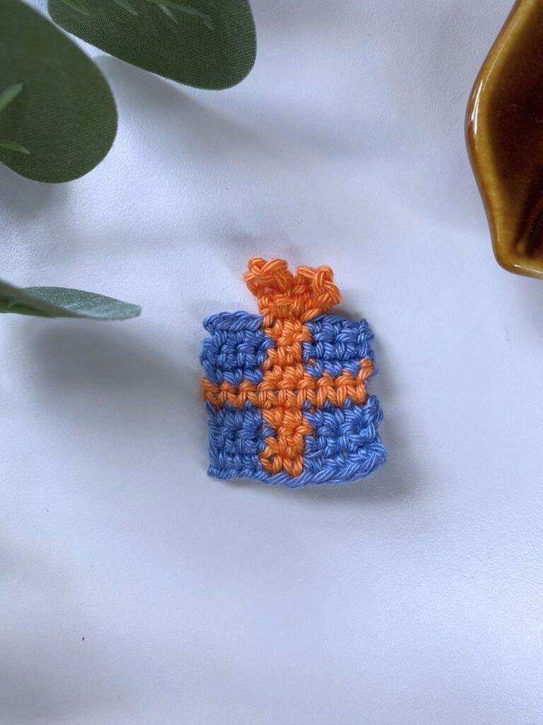 A tiny crochet Christmas present decoration is on a white surface with green and the edge of something brown above