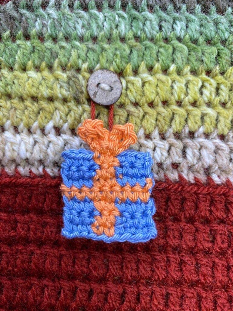 A blue crochet Christmas present ornament with an orange ribbon hangs from a button on a crochet background