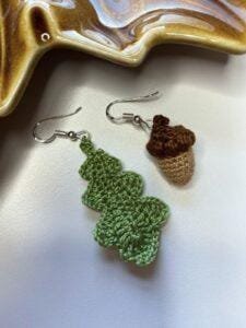 A tiny crochet oak leaf earring and a tiny crochet acorn earring on a white surface in front of a brown leaf shaped dish
