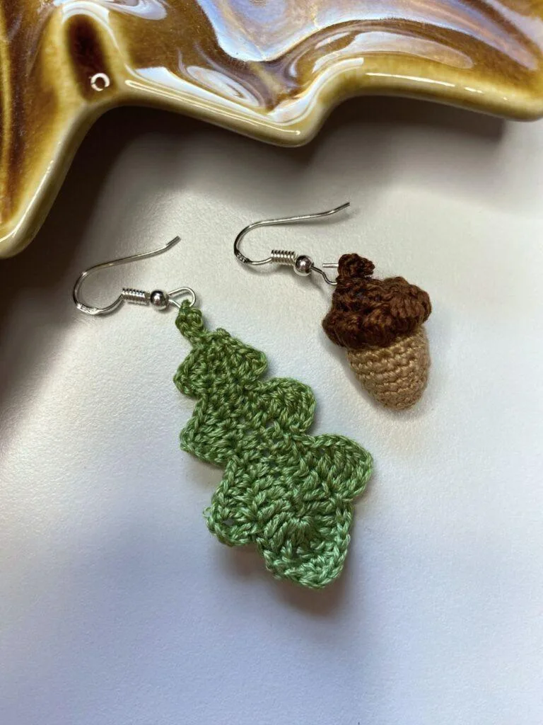 A tiny crochet oak leaf earring and a tiny crochet acorn earring on a white surface in front of a brown leaf shaped dish