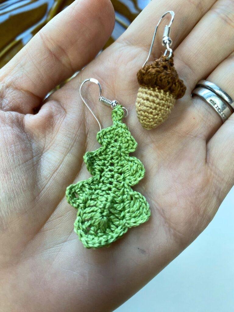 A flat hand shows a pair of micro crochet earrings of an oak leaf and an acorn