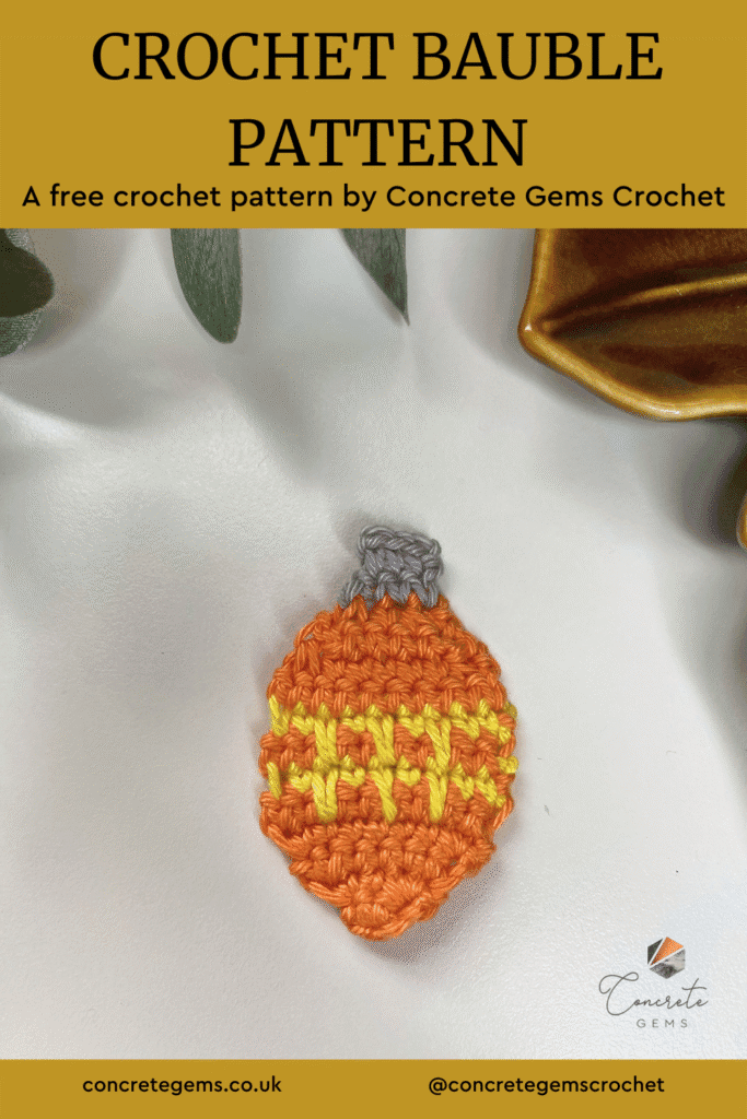 A flat orange crochet bauble with yellow stripes and a silver top lays on a white surface with a brown dish and greenery above and the words CROCHET BAUBLE PATTERN are above