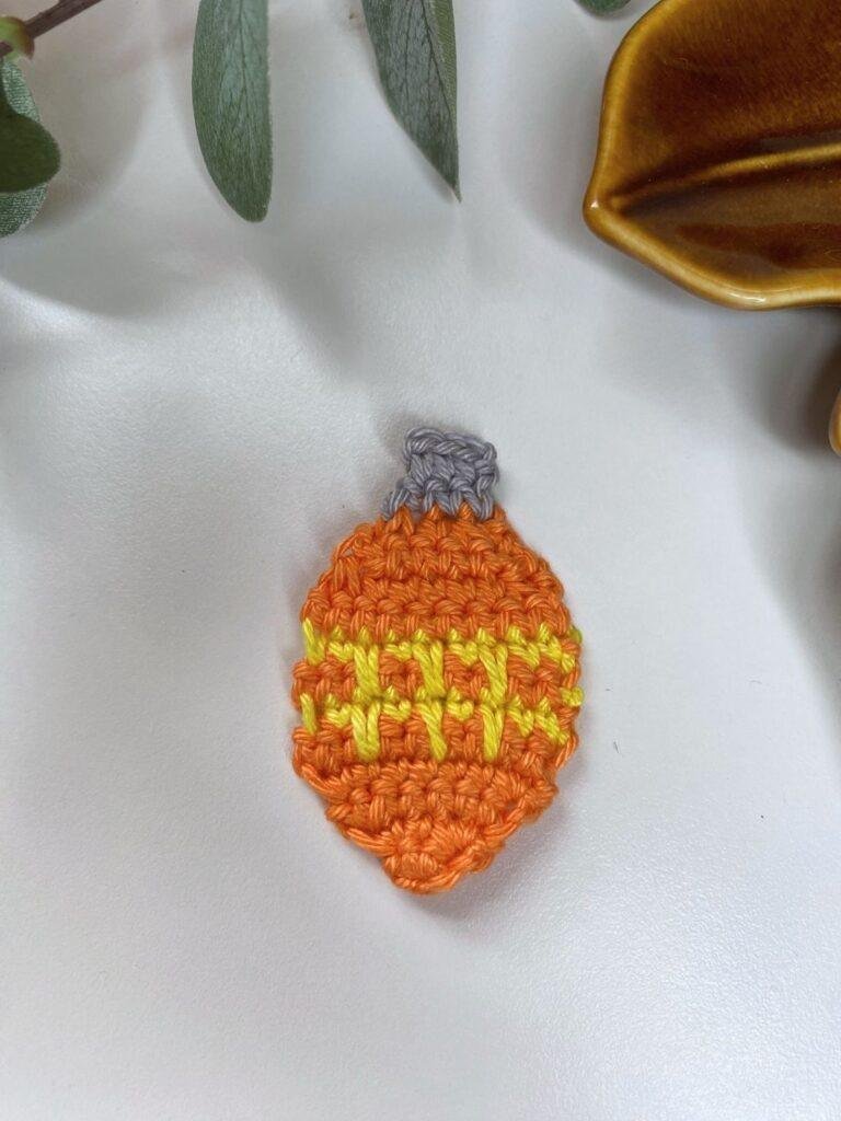 A flat orange crochet bauble with yellow stripes and a silver top lays on a white surface with a brown dish and greenery above