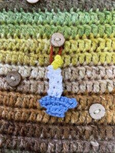 A tiny crochet candle stick hangs from a button on a brown and green crochet background