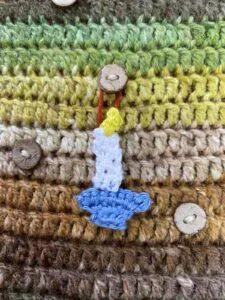 A tiny crochet candle stick hangs from a button on a brown and green crochet background
