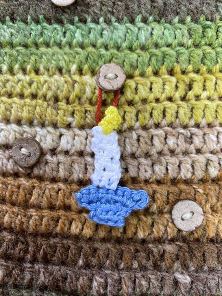 A tiny crochet candle stick hangs from a button on a brown and green crochet background