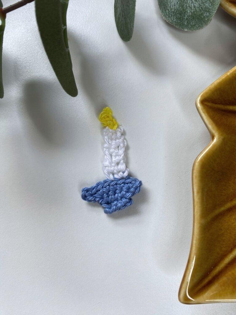 A small crochet candle holder lays on a white background surrounded by greenery and a brown plate
