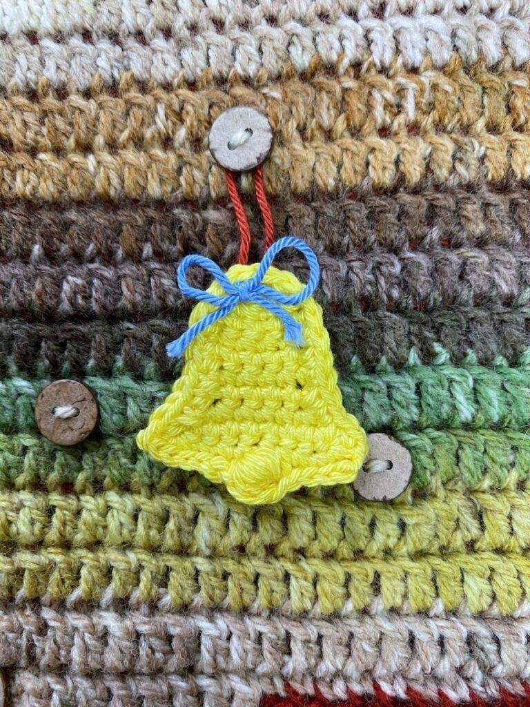 A yellow crochet bell with a blue ribbon hangs on a button on a brown and green crochet background