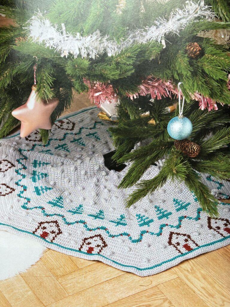 A Christmas tree has pink and silver decorations and a silver crochet Christmas tree skirt underneath with trees and cabins on