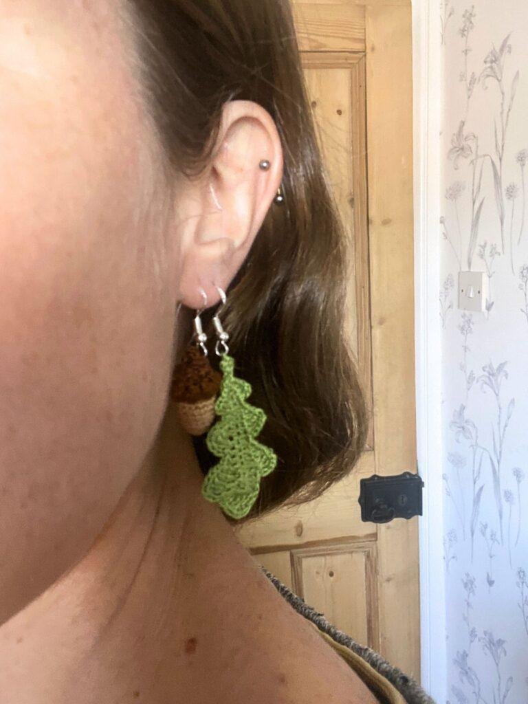 A close up of a person's ear showing them wearing crochet acorn and oak leaf earrings in 2 holes of the same ear