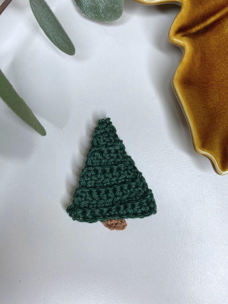 A mini crochet Christmas tree is on a white background with greenery and a brown dish at the edge