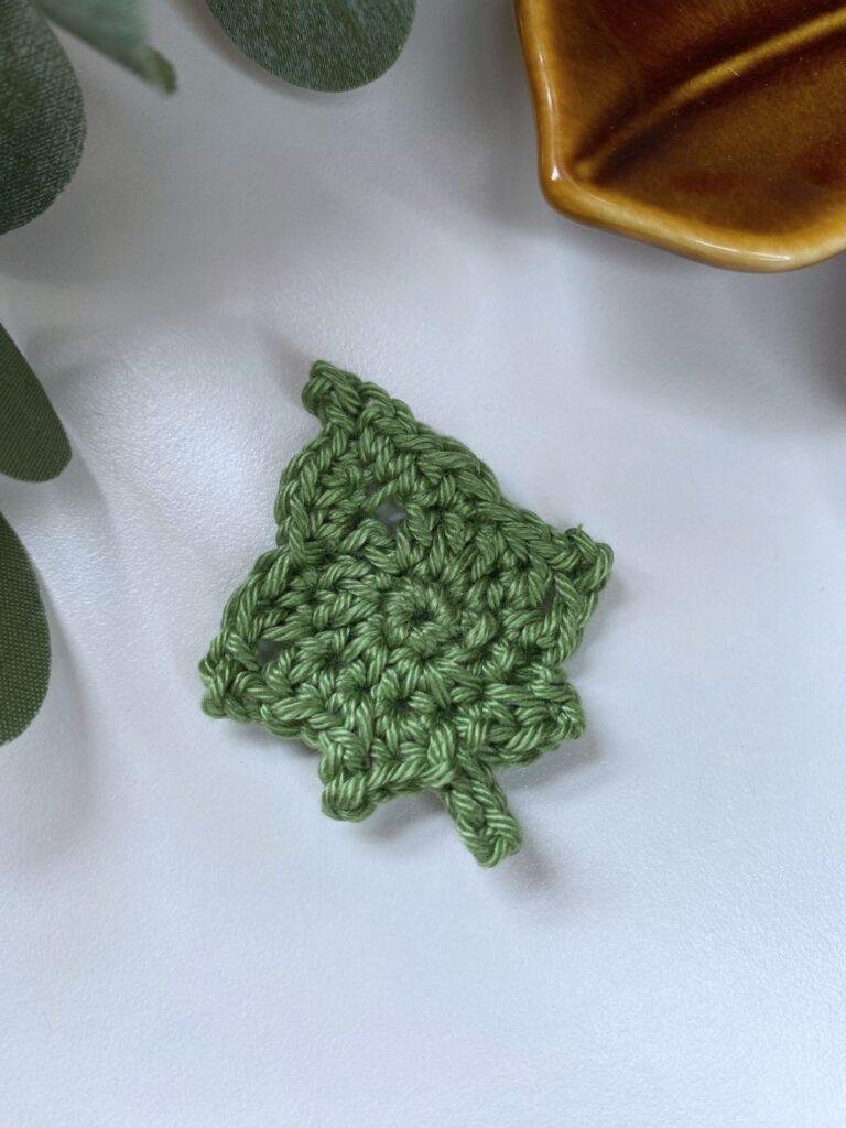 A green crochet ivy leaf lays on a white background with greenery and a brown edge of a dish