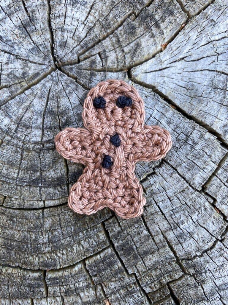 A small crochet gingerbread man ornament lays on a wooden slice