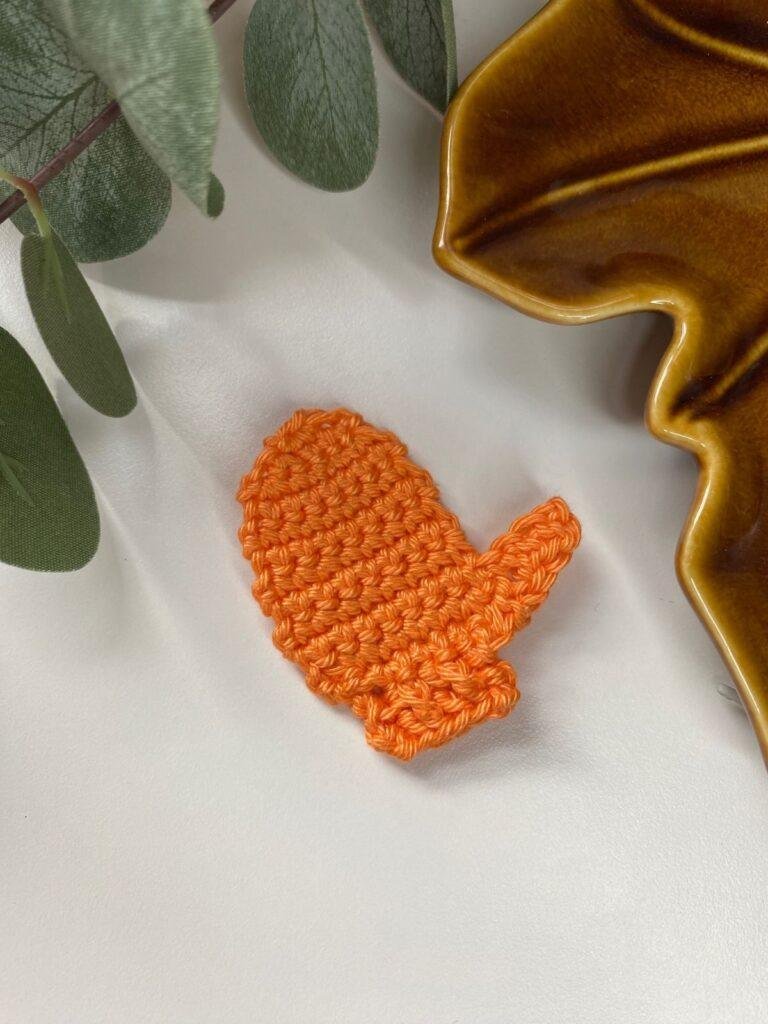 A flat orange crochet mitten decoration lies on a white background with leaves and a brown dish around the edge