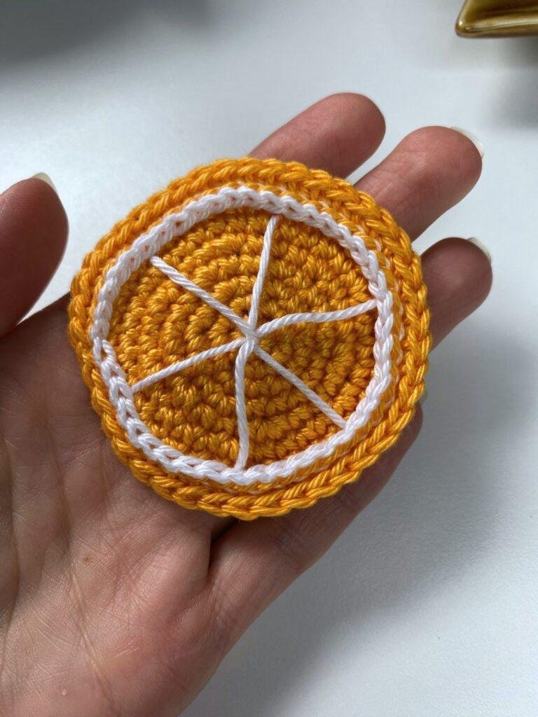 A hadn't holds a crochet orange slice over a white background