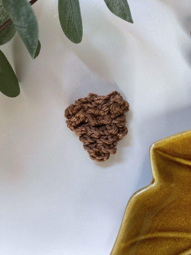 A brown crochet pine cone on a white surface with greenery and brown ceramic in the corners 