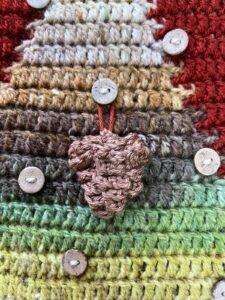 A brown crochet pinecone with lots of texture hangs from a button on a crochet Christmas tree