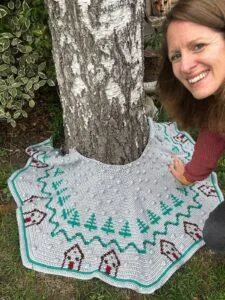 A crochet Christmas tree skirt is wrapped around a tree growing in a garden with a woman's face just leaning in the side of the frame