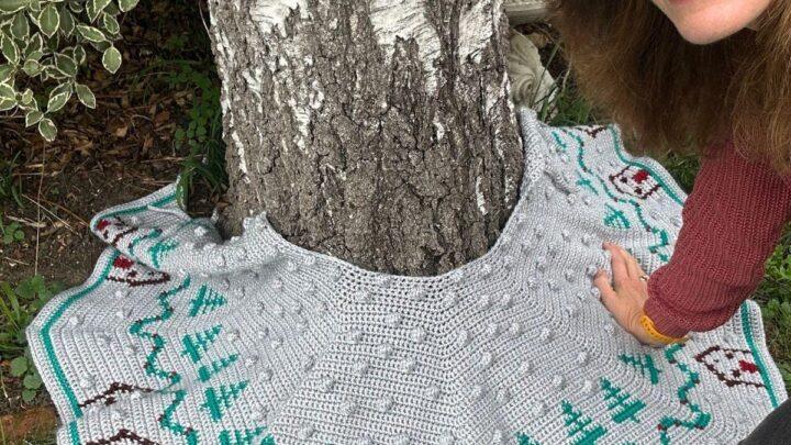 A crochet Christmas tree skirt is wrapped around a tree growing in a garden with a woman's face just leaning in the side of the frame