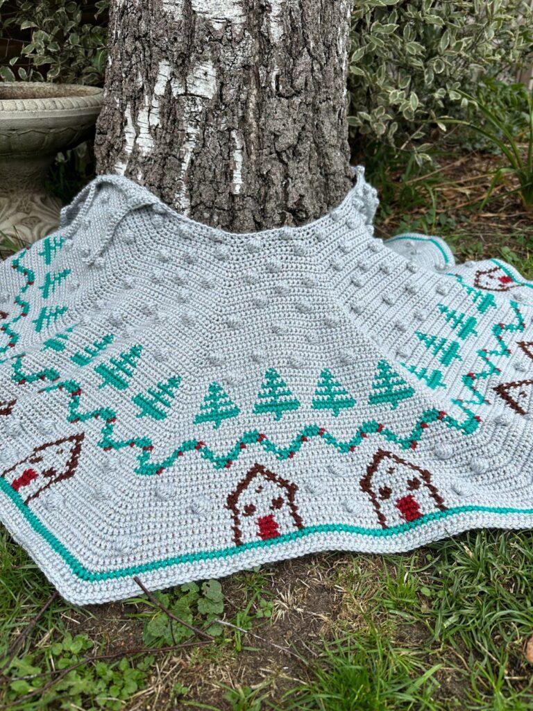 a crochet Christmas tree skirt with bobbles, tree and huts on is draped round a tree in a garden