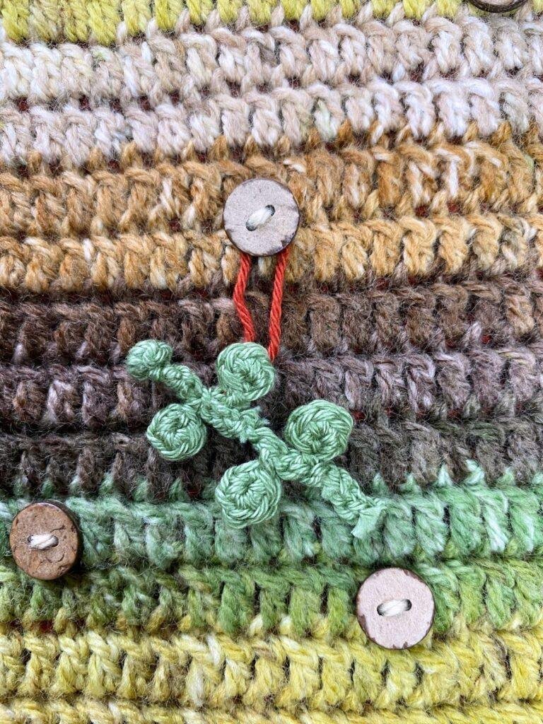 A tiny crochet eucalyptus hangs on a button on a brown and green crochet background