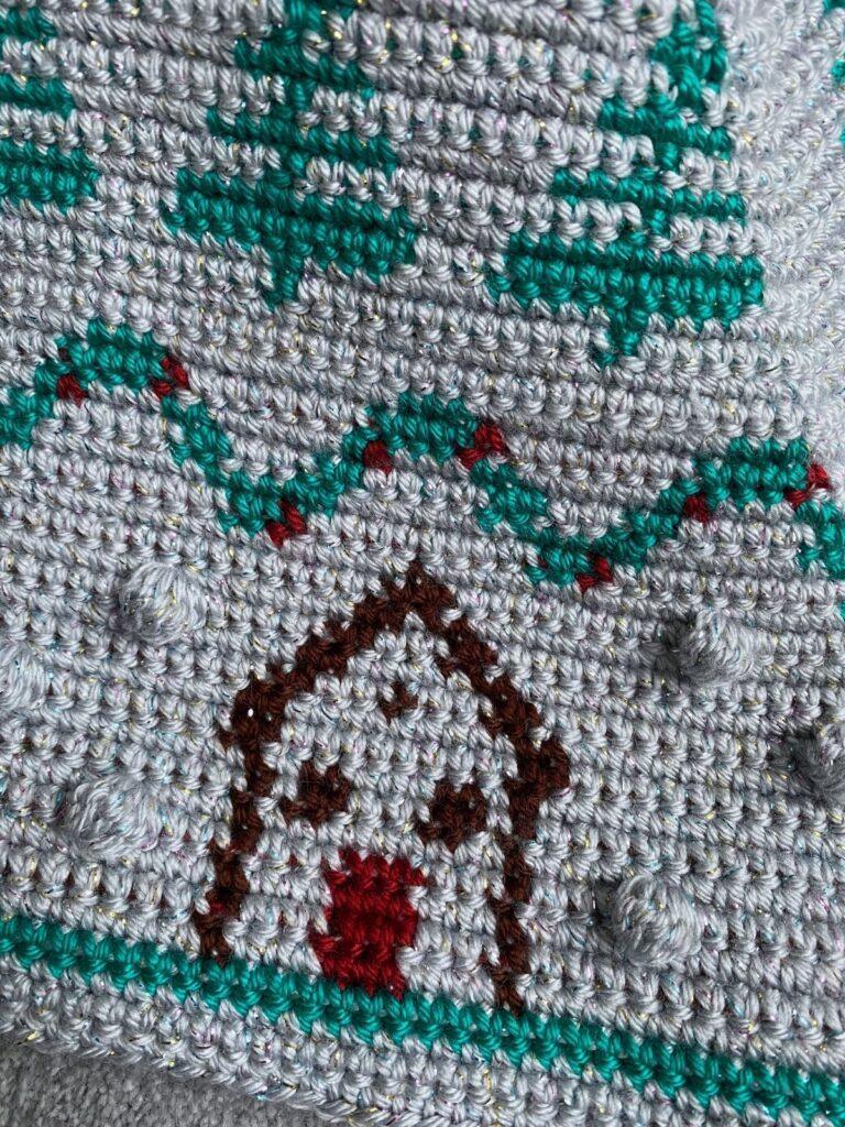 close up of a silver crochet fabric with bobbles on, green trees, a green and red garland and a wooden hut with a red door