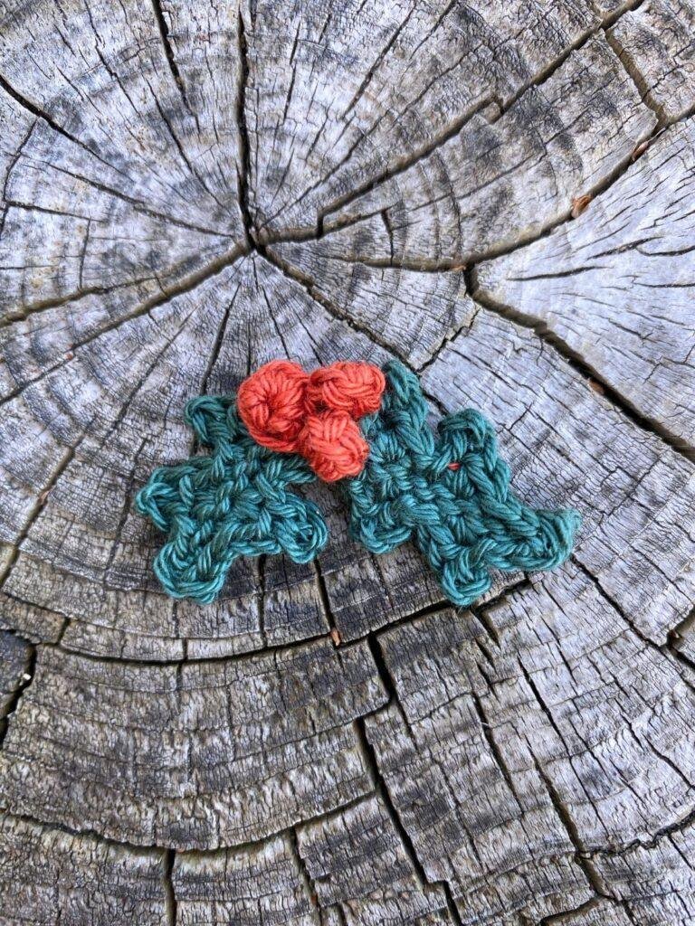 A small crochet holly and berries decoration lays on a wooden slice