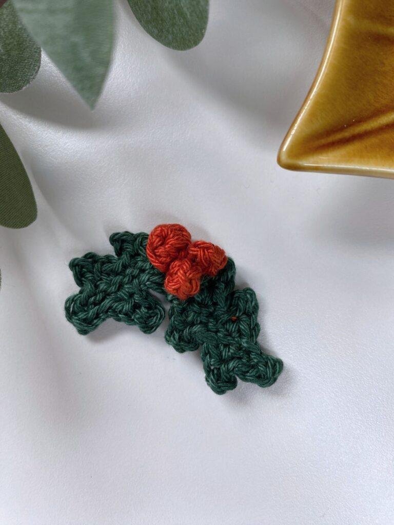 A small crochet holly leaves and berries ornament on a white background with the post of a leaf shaped dish off screen