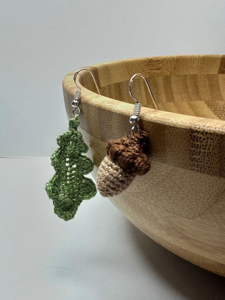 a pair of crochet acorn and oak leaf earrings hang from the edge of a wooden bowl