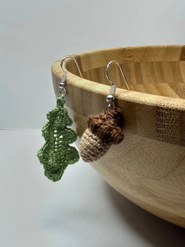 a pair of crochet acorn and oak leaf earrings hang from the edge of a wooden bowl